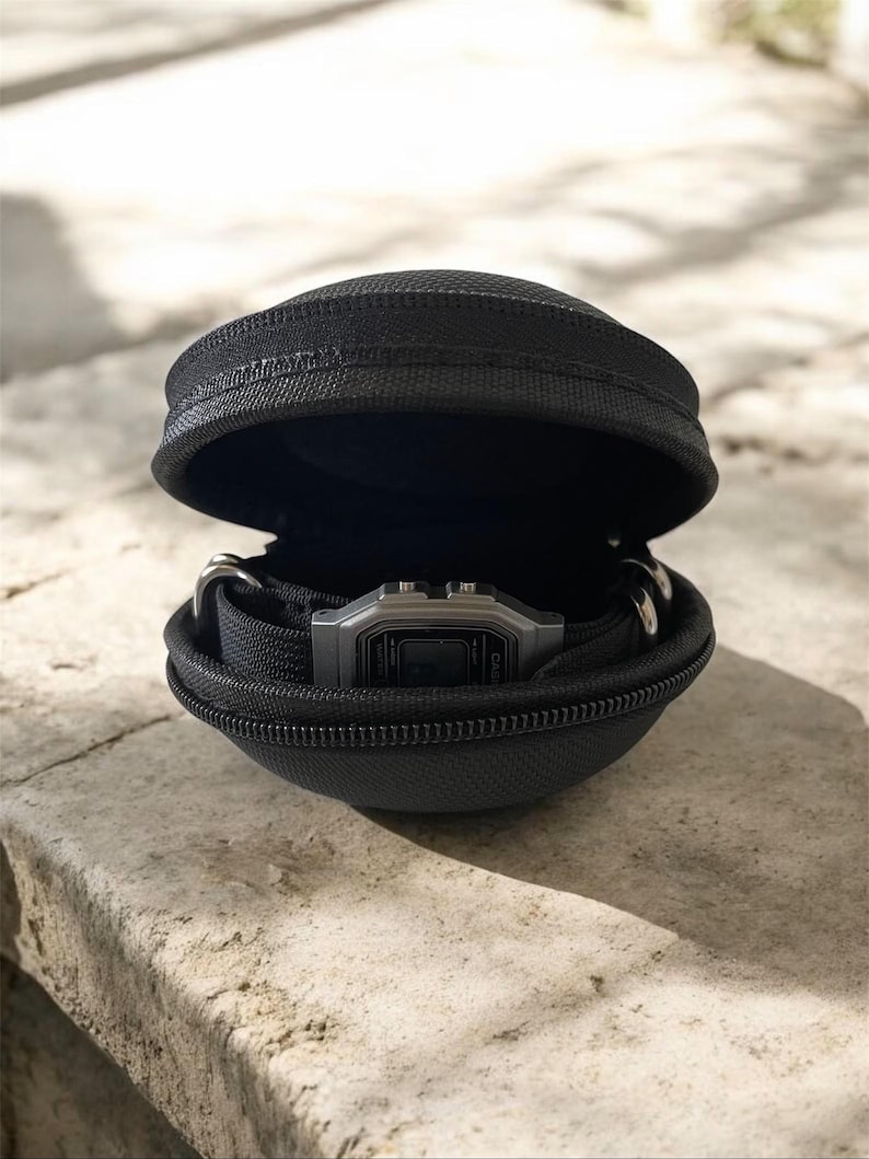 Puede incluir: Un estuche de transporte negro, semirr&iacute;gido, con cremallera y forma de concha que contiene un reloj de pulsera digital con esfera rectangular plateada y correa negra. El estuche est&aacute; entreabierto, mostrando el reloj en su interior.