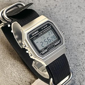 May include: A silver Casio F-91W digital watch with a black nylon strap. The watch face displays the time, day, and date. The watch has a rectangular face and is water-resistant. The strap is secured with silver metal loops.