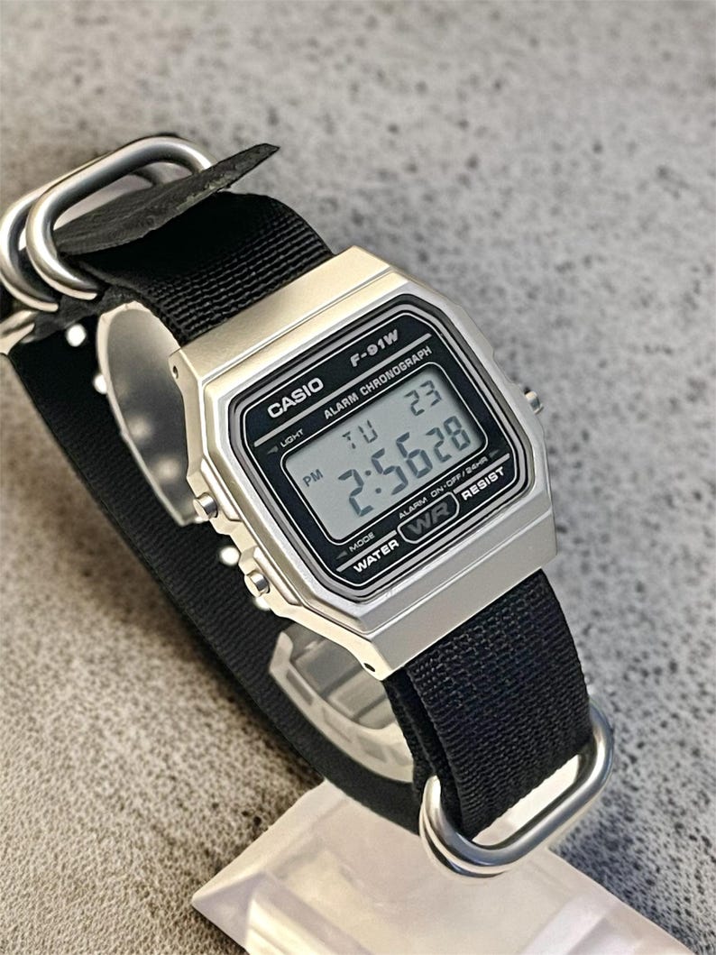 May include: A silver Casio F-91W digital watch with a black nylon strap. The watch face displays the time, day, and date. The watch has a rectangular face and is water-resistant. The strap is secured with silver metal loops.