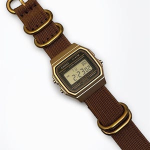 Może przedstawiać: Cyfrowy zegarek Casio F-91W z brązowym nylonowym paskiem i złotymi elementami. Tarcza zegarka wyświetla godzinę, dzień i datę. Zegarek ma napis "CASIO", "F-91W", "ALARM CHRONOGRAPH" i "WATER WR RESIST".