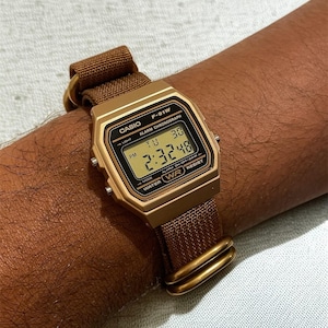 Może przedstawiać: Złoty cyfrowy zegarek Casio F-91W z brązowym paskiem z tkaniny. Tarcza zegarka wyświetla godzinę, dzień i datę. Zegarek jest przymocowany do nadgarstka za pomocą brązowego paska i złotych element&oacute;w.