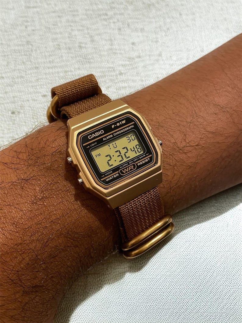 Może przedstawiać: Złoty cyfrowy zegarek Casio F-91W z brązowym paskiem z tkaniny. Tarcza zegarka wyświetla godzinę, dzień i datę. Zegarek jest przymocowany do nadgarstka za pomocą brązowego paska i złotych element&oacute;w.