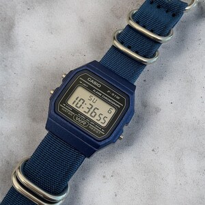Puede incluir: Un reloj digital azul con esfera rectangular y correa de nailon azul a juego con herrajes de metal plateado. La pantalla muestra el texto CASIO, F-91W, ALARM CHRONOGRAPH, WATER WR RESIST y la hora 10:36:55 con SU 6.