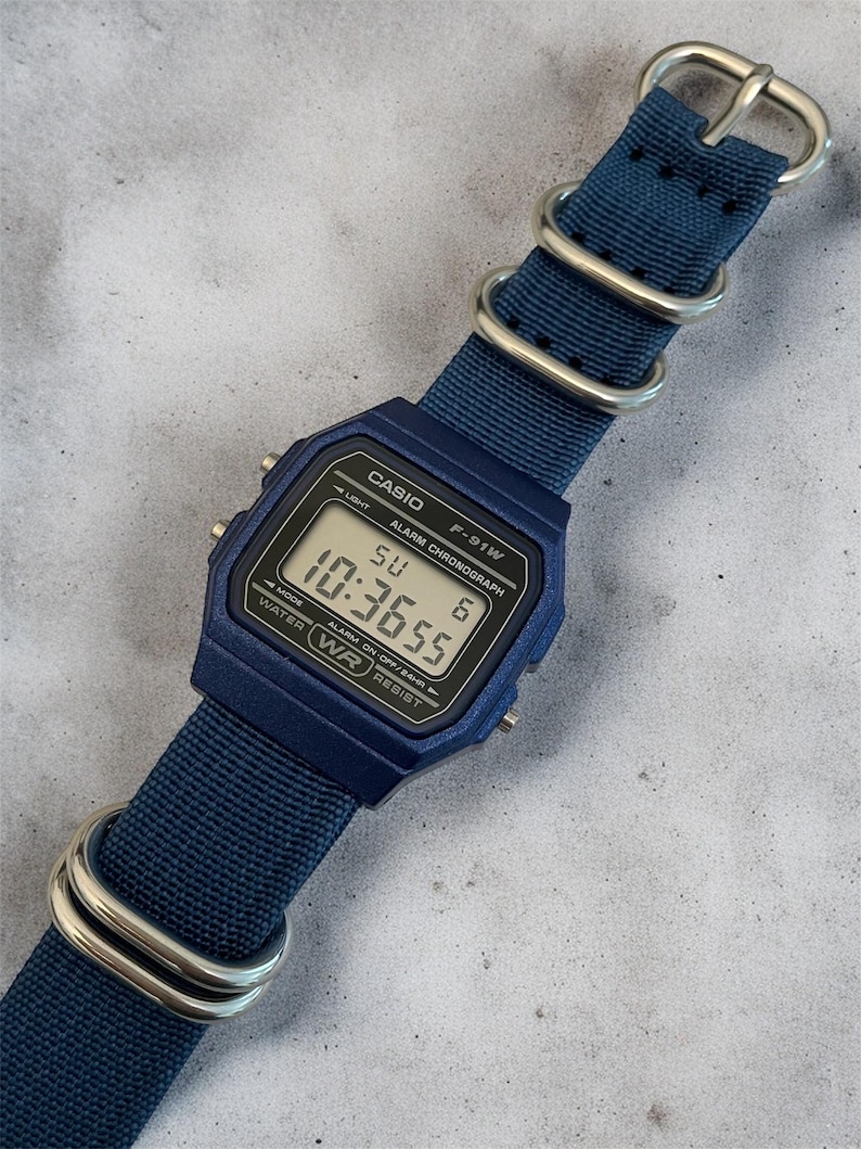 Puede incluir: Un reloj digital azul con esfera rectangular y correa de nailon azul a juego con herrajes de metal plateado. La pantalla muestra el texto CASIO, F-91W, ALARM CHRONOGRAPH, WATER WR RESIST y la hora 10:36:55 con SU 6.