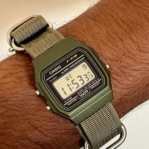 May include: A digital Casio F-91W watch with a green case and a green fabric strap. The watch face displays the time as 11:53:36. The watch has a water-resistant feature and is secured with a silver-tone metal buckle.