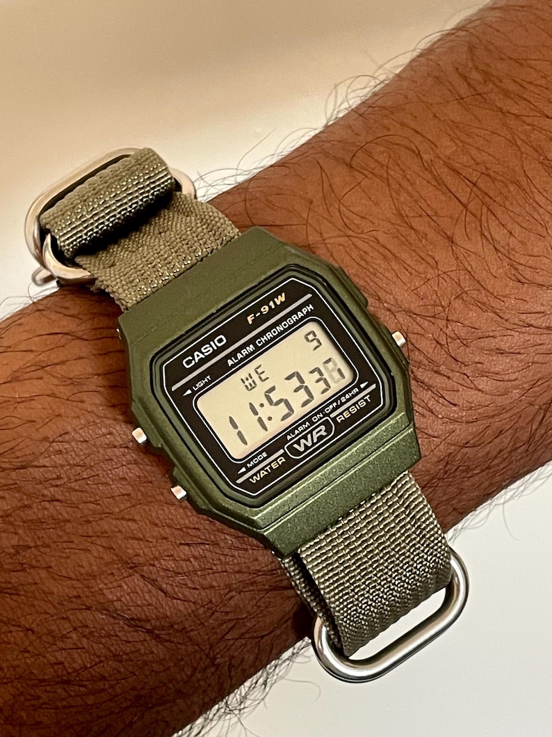 May include: A digital Casio F-91W watch with a green case and a green fabric strap. The watch face displays the time as 11:53:36. The watch has a water-resistant feature and is secured with a silver-tone metal buckle.
