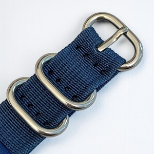 Puede incluir: Una correa de reloj de nailon azul marino con textura tejida y una serie de orificios de ajuste termosellados. La correa se asegura con tres anillos de metal plateado pulido, que incluyen una hebilla y dos trabillas rectangulares.
