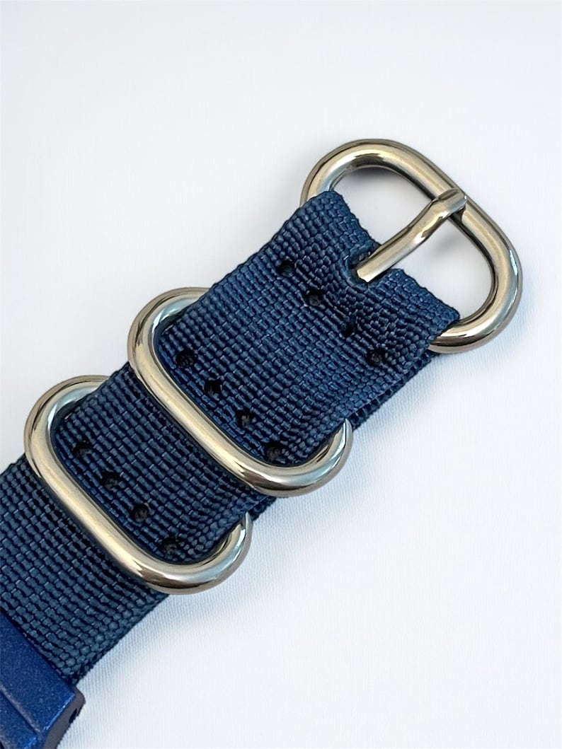 Puede incluir: Una correa de reloj de nailon azul marino con textura tejida y una serie de orificios de ajuste termosellados. La correa se asegura con tres anillos de metal plateado pulido, que incluyen una hebilla y dos trabillas rectangulares.