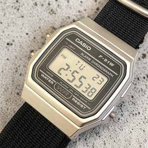 May include: A silver Casio F-91W digital watch with a black nylon strap. The watch face displays the time, day, and date. The watch has the text "CASIO", "F-91W", and "WATER RESIST" printed on the face.