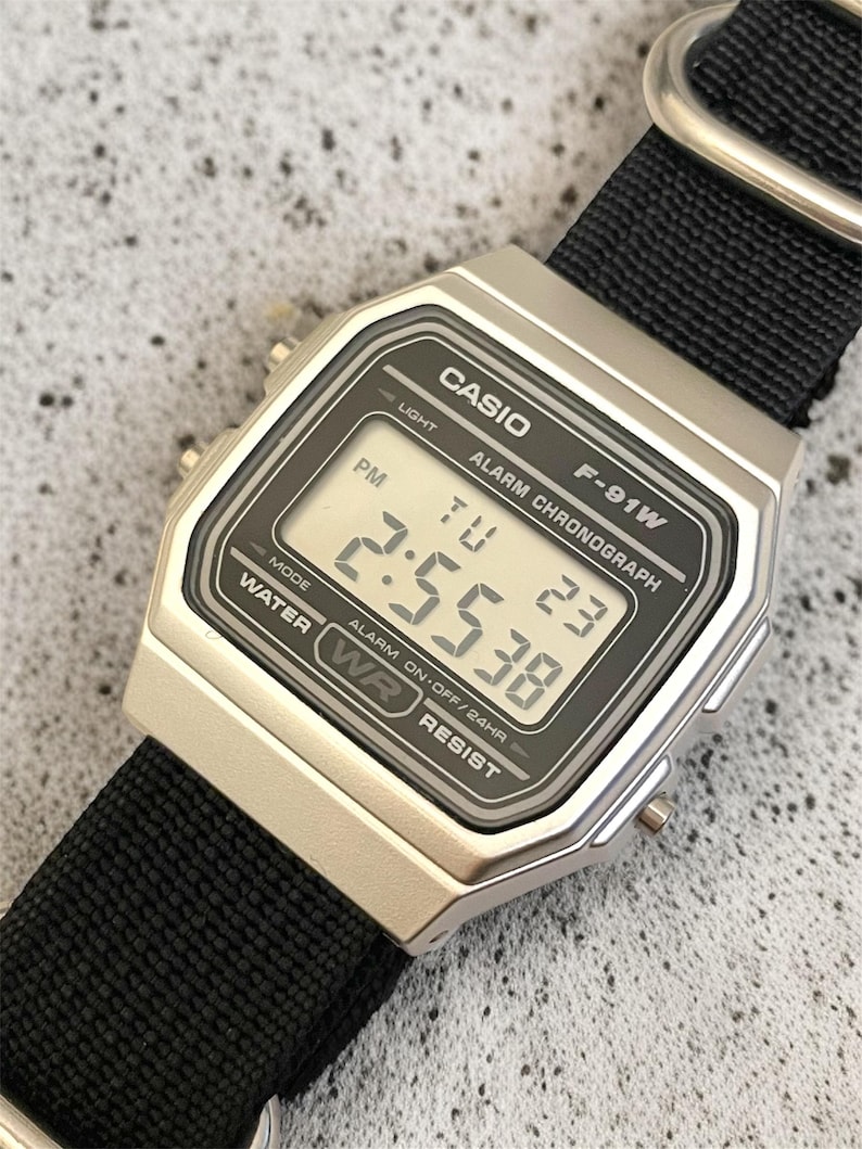 May include: A silver Casio F-91W digital watch with a black nylon strap. The watch face displays the time, day, and date. The watch has the text "CASIO", "F-91W", and "WATER RESIST" printed on the face.