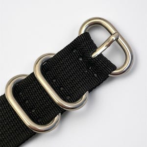 May include: Close-up of a black watch strap with silver-colored metal hardware. The strap is made of a woven material and features two metal loops and a buckle. The hardware is shiny and contrasts with the dark strap.