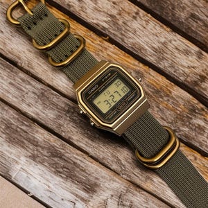 May include: A digital Casio F-91W watch with a gold-tone case and olive green nylon strap. The watch displays the time and date. The strap features gold-colored metal hardware. The watch is set against a wooden background.