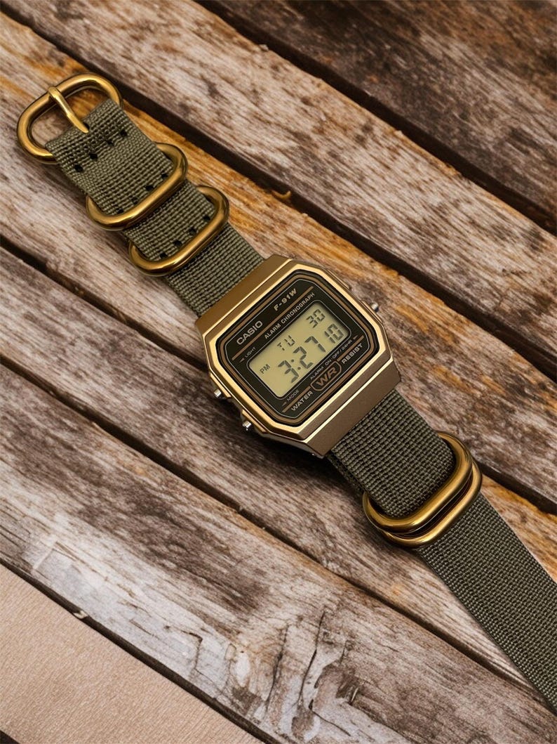 May include: A digital Casio F-91W watch with a gold-tone case and olive green nylon strap. The watch displays the time and date. The strap features gold-colored metal hardware. The watch is set against a wooden background.