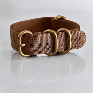 Puede incluir: Una correa de reloj marrón con herrajes metálicos dorados. La correa está hecha de un material tejido y presenta una hebilla y varios bucles. La correa del reloj está diseñada para ser duradera y elegante.