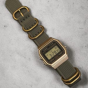 May include: A gold-toned Casio F-91W digital watch with a green nylon strap and gold-colored hardware. The watch face displays the time, day, and date. The strap is secured with a buckle and two loops.