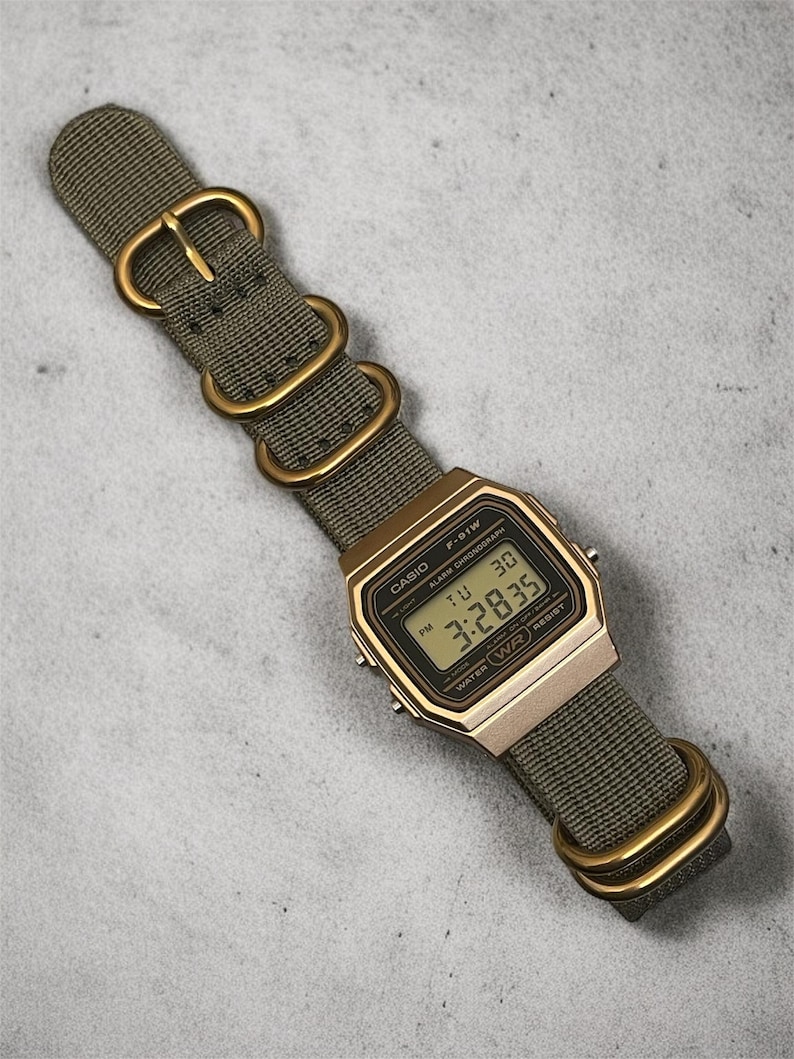 May include: A gold-toned Casio F-91W digital watch with a green nylon strap and gold-colored hardware. The watch face displays the time, day, and date. The strap is secured with a buckle and two loops.