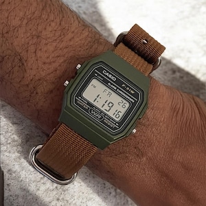 May include: A digital Casio F-91W watch with a green case and a brown fabric strap. The watch face displays the time as 1:19:16 PM and the date as FR 26. The watch has a light, alarm, and chronograph functions.