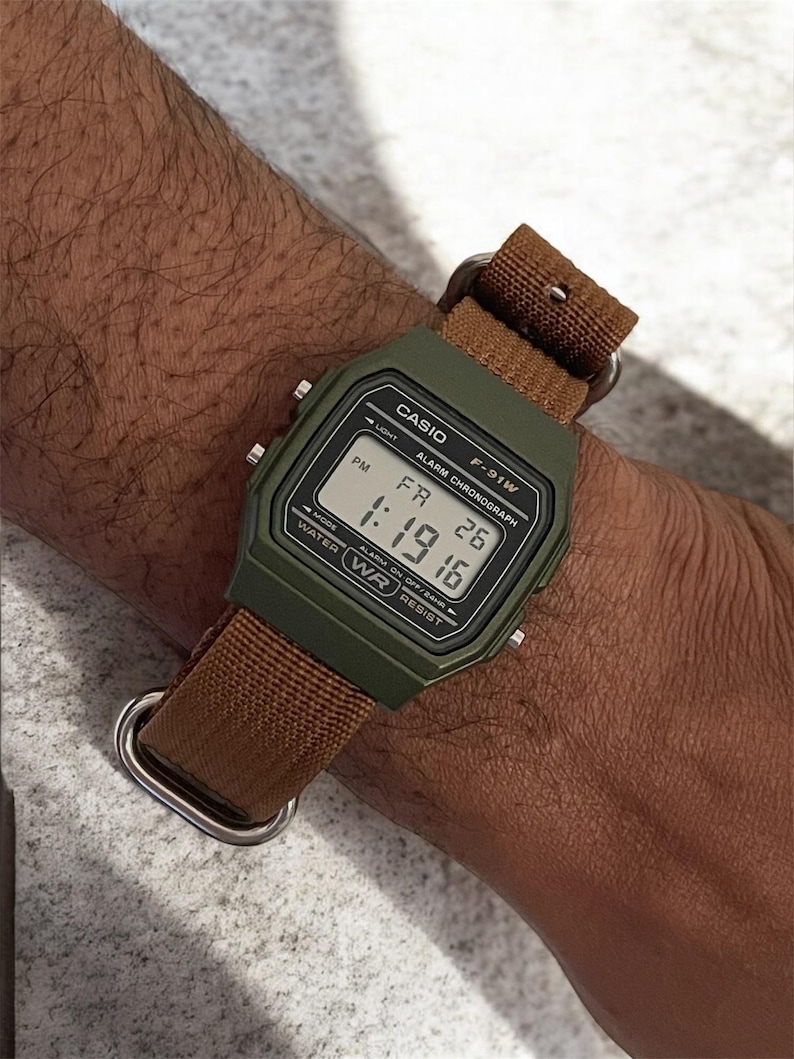 May include: A digital Casio F-91W watch with a green case and a brown fabric strap. The watch face displays the time as 1:19:16 PM and the date as FR 26. The watch has a light, alarm, and chronograph functions.