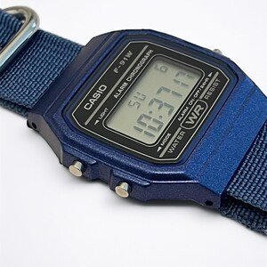 Puede incluir: Un reloj digital azul oscuro con esfera rectangular y correa de tela a juego. La pantalla muestra el texto CASIO, F-91W, ALARM CHRONOGRAPH y WATER RESIST. La pantalla digital indica la hora y el d&iacute;a, con botones laterales plateados.