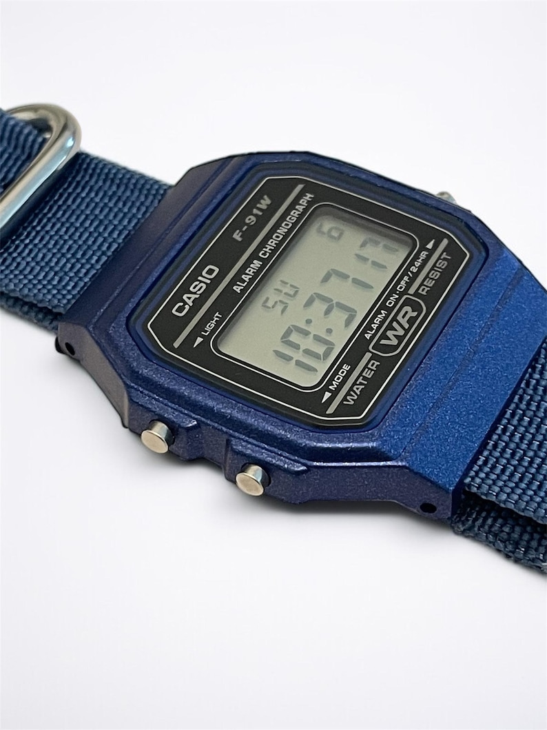 Puede incluir: Un reloj digital azul oscuro con esfera rectangular y correa de tela a juego. La pantalla muestra el texto CASIO, F-91W, ALARM CHRONOGRAPH y WATER RESIST. La pantalla digital indica la hora y el d&iacute;a, con botones laterales plateados.