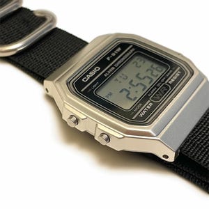 May include: A silver Casio F-91W digital watch with a black nylon strap. The watch face displays the time as 2:55:26 PM. The watch has a rectangular face with the Casio logo and other text.