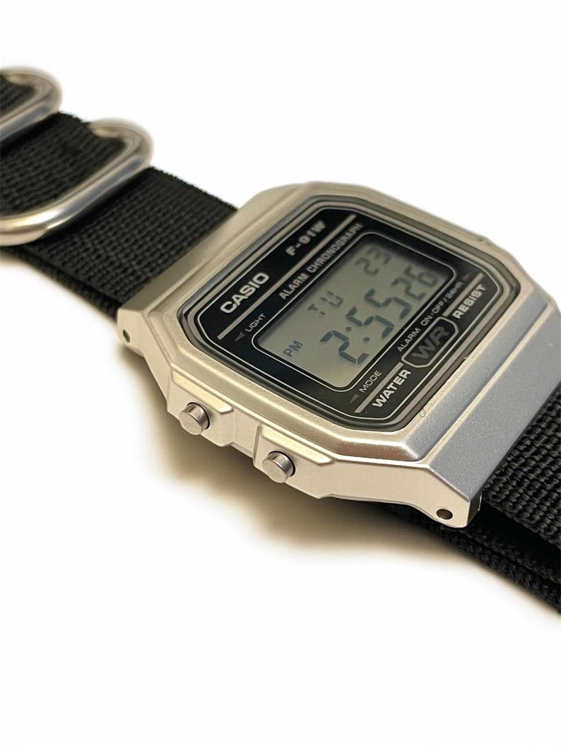 May include: A silver Casio F-91W digital watch with a black nylon strap. The watch face displays the time as 2:55:26 PM. The watch has a rectangular face with the Casio logo and other text.