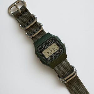 May include: A digital Casio F-91W watch with a dark green case and a green nylon strap. The watch face displays the time, day, and date. The strap has silver-tone metal hardware.
