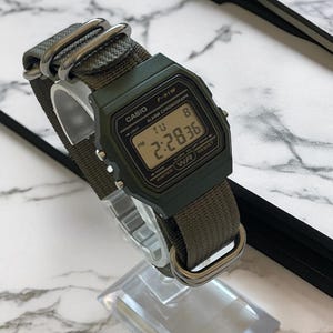 May include: A digital Casio F-91W watch with a green face and olive green nylon strap. The watch displays the time 2:28:36. The watch has the text "CASIO", "F-91W", "ALARM CHRONOGRAPH", and "WATER RESIST" printed on the face.