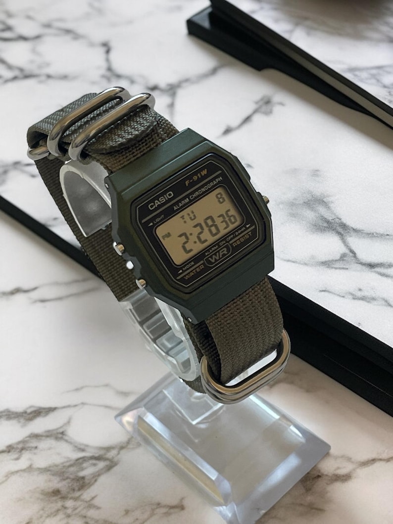 May include: A digital Casio F-91W watch with a green face and olive green nylon strap. The watch displays the time 2:28:36. The watch has the text "CASIO", "F-91W", "ALARM CHRONOGRAPH", and "WATER RESIST" printed on the face.