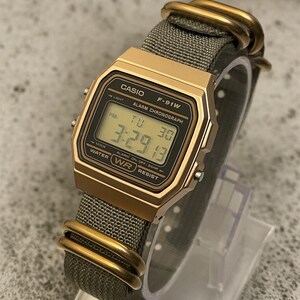 May include: A gold-toned Casio F-91W digital watch with a gray fabric band. The watch face displays the time, day, and date. The band is secured with gold-colored metal loops.
