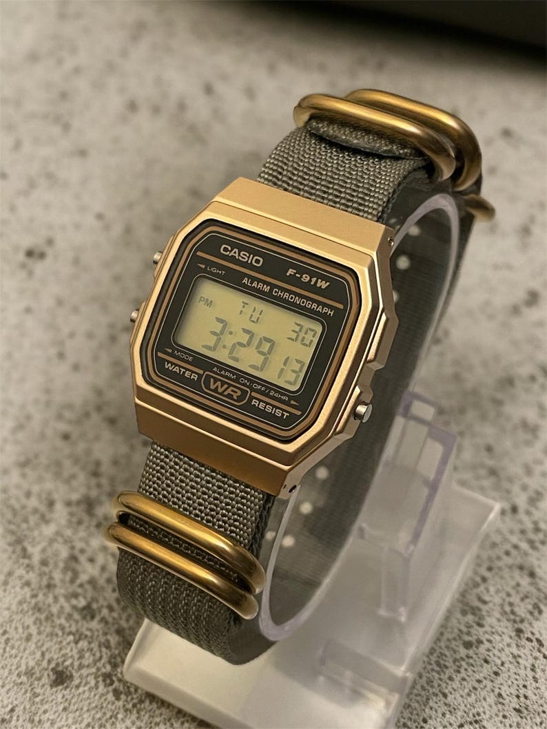 May include: A gold-toned Casio F-91W digital watch with a gray fabric band. The watch face displays the time, day, and date. The band is secured with gold-colored metal loops.