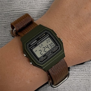 May include: A Casio F-91W digital watch with a green case and a brown nylon strap. The watch face displays the time, day, and date. The watch is secured on a wrist with a brown strap and silver hardware.