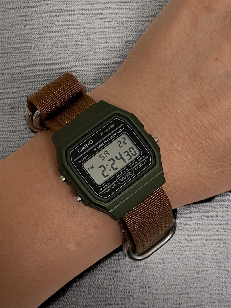 May include: A Casio F-91W digital watch with a green case and a brown nylon strap. The watch face displays the time, day, and date. The watch is secured on a wrist with a brown strap and silver hardware.