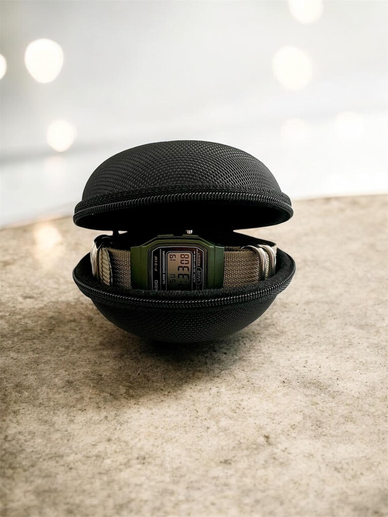 May include: A digital watch with a green face and a beige band is displayed inside a black, zippered, spherical case. The watch shows the time. The case is open, revealing the watch.