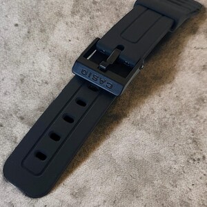 Puede incluir: Una correa de reloj negra mate hecha de material de resina flexible. La correa presenta una serie de orificios de ajuste rectangulares y una hebilla negra a juego con el nombre de la marca CASIO grabado en letras may&uacute;sculas en la hebilla.