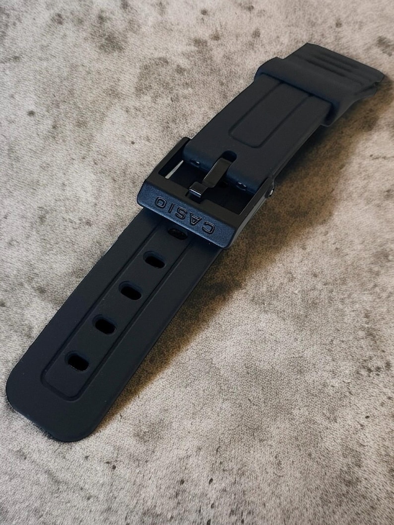 Puede incluir: Una correa de reloj negra mate hecha de material de resina flexible. La correa presenta una serie de orificios de ajuste rectangulares y una hebilla negra a juego con el nombre de la marca CASIO grabado en letras may&uacute;sculas en la hebilla.