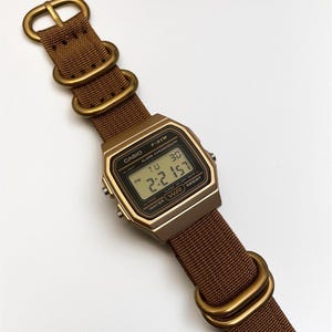 Może przedstawiać: Cyfrowy zegarek Casio F-91W z brązowym paskiem i złotymi elementami. Tarcza zegarka wyświetla godzinę, datę i inne funkcje. Pasek wykonany jest z tkanego materiału.
