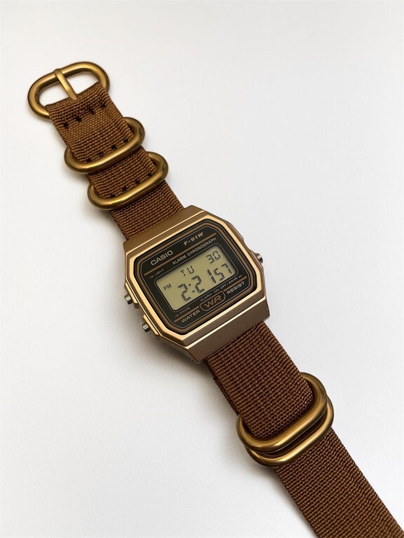 Może przedstawiać: Cyfrowy zegarek Casio F-91W z brązowym paskiem i złotymi elementami. Tarcza zegarka wyświetla godzinę, datę i inne funkcje. Pasek wykonany jest z tkanego materiału.