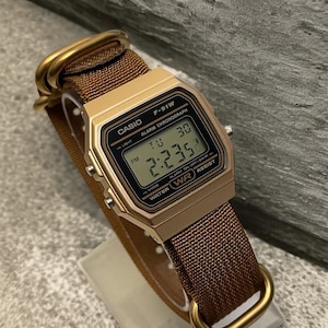 Może przedstawiać: Złoty zegarek cyfrowy Casio F-91W z brązowym nylonowym paskiem. Tarcza zegarka wyświetla godzinę, dzień i datę. Zegarek ma prostokątną kopertę i złote metalowe elementy. Pasek jest zabezpieczony złotymi metalowymi pętlami.