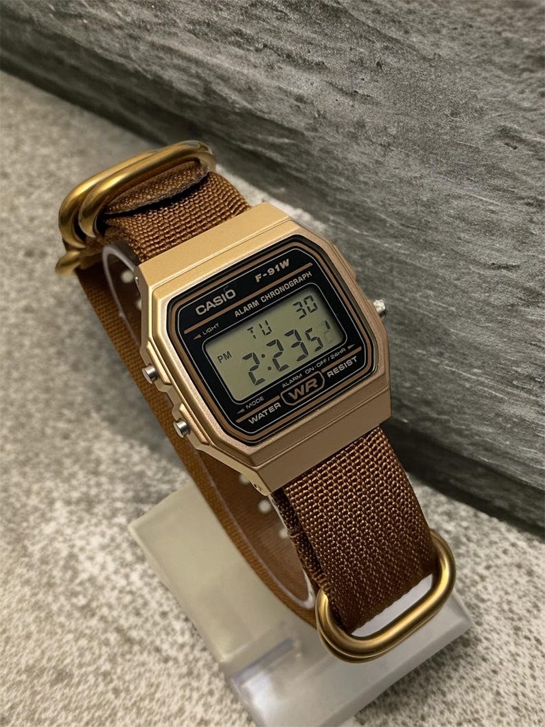 Może przedstawiać: Złoty zegarek cyfrowy Casio F-91W z brązowym nylonowym paskiem. Tarcza zegarka wyświetla godzinę, dzień i datę. Zegarek ma prostokątną kopertę i złote metalowe elementy. Pasek jest zabezpieczony złotymi metalowymi pętlami.