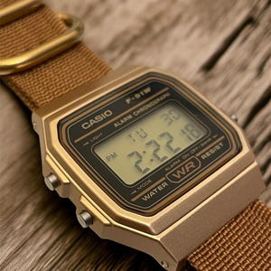 Może przedstawiać: Złoty zegarek cyfrowy Casio F-91W z brązowym nylonowym paskiem. Tarcza zegarka pokazuje godzinę 14:22 i datę. Zegarek ma prostokątną kopertę i posiada napisy "CASIO" i "WATER RESIST".
