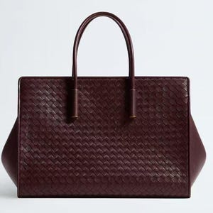 Puede incluir: Bolso tote de cuero burdeos con un patrón tejido. El bolso tiene dos asas arqueadas y una forma rectangular con lados ligeramente inclinados. Las asas están unidas con herrajes dorados.