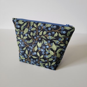 Puede incluir: Una pequeña bolsa trapezoidal con cremallera azul. La tela tiene un fondo azul oscuro y un patrón repetido de arándanos, hojas verdes y tallos marrones. La bolsa es probablemente para cosméticos o artículos pequeños.