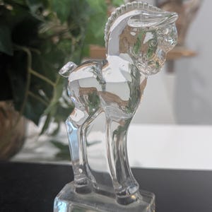 May include: A clear glass figurine of a horse standing on a rectangular base. The horse has detailed features, including a mane and tail. The figurine is transparent, allowing light to pass through, and is likely a decorative item.