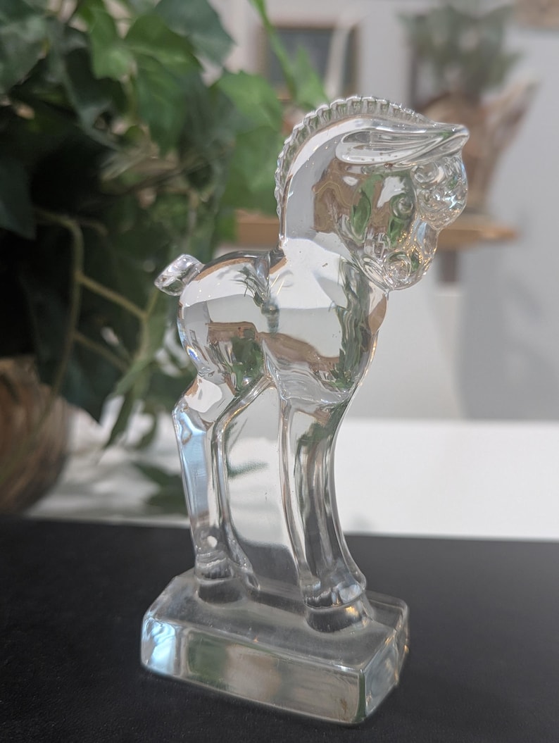 May include: A clear glass figurine of a horse standing on a rectangular base. The horse has detailed features, including a mane and tail. The figurine is transparent, allowing light to pass through, and is likely a decorative item.