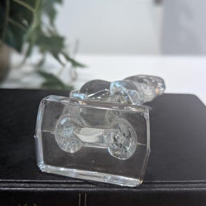 May include: A clear glass figurine of a reclining animal, possibly a cat or dog, atop a rectangular base. The base has two circular indentations. The figurine is on a dark surface, with a blurred background of greenery and a white wall.