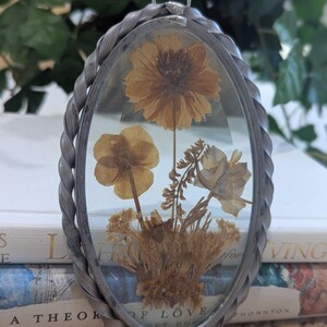 May include: Oval pendant with dried flowers encased in clear glass. The pendant has a silver-colored twisted metal frame and a small loop at the top for hanging. The flowers are various shades of brown and yellow.