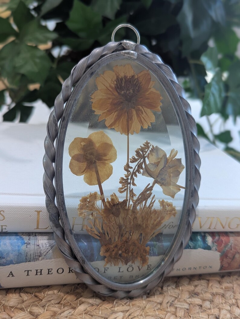 May include: Oval pendant with dried flowers encased in clear glass. The pendant has a silver-colored twisted metal frame and a small loop at the top for hanging. The flowers are various shades of brown and yellow.