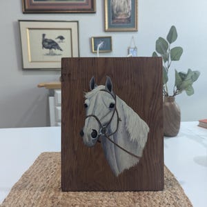 May include: A wooden plaque featuring a painted portrait of a white horse's head with a bridle. The horse is set against a dark brown wood grain background. The artwork is displayed on a woven jute mat.