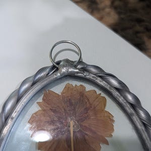 May include: A pressed flower art piece in a silver-colored frame. The frame has a braided design around the edges and a small ring at the top for hanging. The flower is a dried, light brown color.